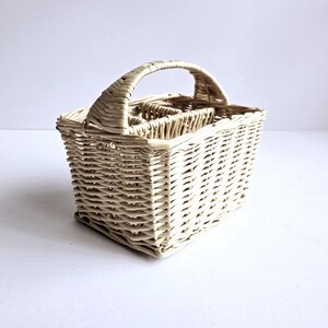 May include: A rectangular, light-colored woven wicker basket featuring a single arched handle attached to the top. The basket has a structured design with an open interior divided into multiple compartments, showcasing a natural, light beige finish.