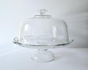 May include: A clear glass cake stand with a dome lid. The stand has a scalloped edge and a pedestal base.