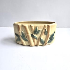 May include: An oval ceramic planter with a cream-colored crackle glaze finish. The exterior features a raised, molded relief design of bamboo stalks and leaves in a muted sage green color, wrapping around the curved sides of the vessel.