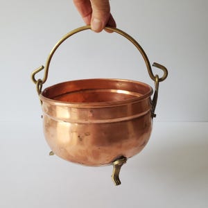 May include: A small copper pot with a brass handle and three brass feet. The pot is round and has a slightly flared rim.