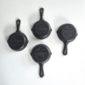 May include: Four small black cast iron skillets arranged in a group. Each skillet features a round cooking surface, a short handle with a hole for hanging, and the embossed text LODGE USA MS on the bottom. The pans have a matte finish.
