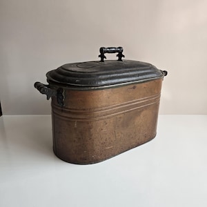May include: An antique copper and black metal food storage container with a hinged lid and a wooden handle. The container has a rounded oval shape and is slightly tarnished.