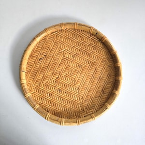 May include: A round, woven bamboo tray with a herringbone pattern. The tray is light brown with a natural finish and a slightly raised rim. The tray is approximately 12 inches in diameter.
