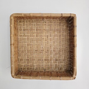 May include: A square, open-top basket constructed from light brown woven reeds. The basket features a structured frame with a grid-like weave pattern on the base and sides, showcasing natural variations in the color and texture of the reed material.