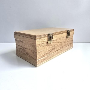 May include: A rectangular wooden box with a light brown wood grain finish. The box features a hinged lid secured by two metallic gold-colored latches on the front. The construction shows visible layered plywood edges along the sides and top lid.