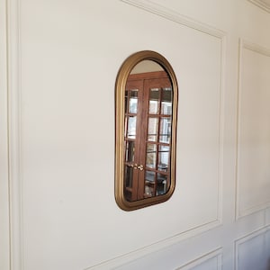 May include: A gold-framed arched mirror hangs on a white wall. The mirror reflects a wooden door with glass panels.