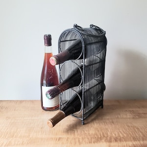 May include: A black wire wine rack with a cylindrical frame and a top handle, holding three glass wine bottles horizontally. One additional wine bottle with a light red liquid and a white label stands upright next to the wire rack structure.