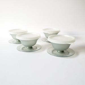 May include: Four vintage gray sundae dishes with white lids and matching saucers. The dishes have a classic, stemmed design. The lids are round and fit snugly on top of the bowls. The saucers are flat and circular, providing a stable base for the dishes.