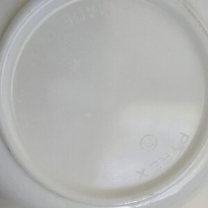May include: A white circular glass dish with a smooth, opaque finish. The underside features embossed text reading PYREX with a registered trademark symbol and the word MADE, visible in a circular arrangement around the center of the base.
