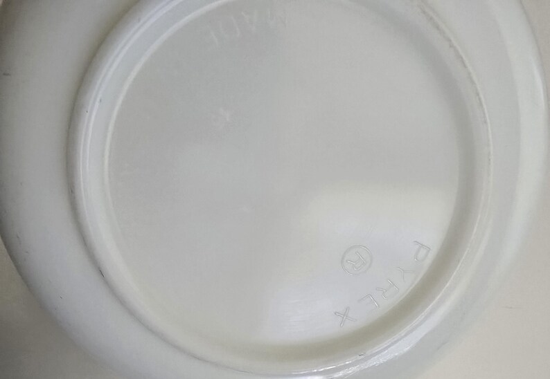 May include: A white circular glass dish with a smooth, opaque finish. The underside features embossed text reading PYREX with a registered trademark symbol and the word MADE, visible in a circular arrangement around the center of the base.