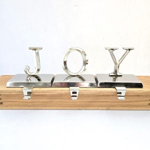 May include: Silver metal Christmas stocking holders spelling out "JOY". Each letter sits atop a square base with a hook for hanging stockings. The holders are mounted on a light-colored wooden base.