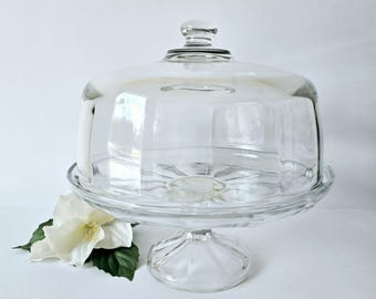 May include: A clear glass cake stand with a domed lid. The stand has a fluted base and a scalloped edge.