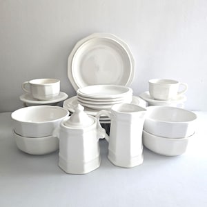 May include: A collection of white ceramic dishware, including plates, bowls, cups, saucers, a sugar bowl, and a creamer. The plates have a scalloped edge design. The set is arranged on a white surface, ideal for serving meals.