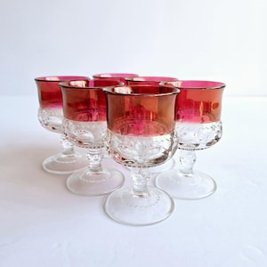 May include: Set of six vintage wine glasses. Each glass features a clear, patterned base and a vibrant red bowl with a gold rim. The glasses are arranged on a white surface, showcasing their elegant design and color contrast.