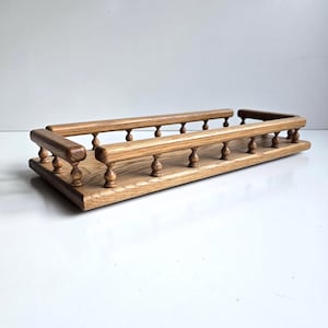 May include: A rectangular wooden tray featuring a natural light brown wood grain finish. The tray is surrounded by a decorative railing supported by small, turned wooden spindles along the front and sides, creating a structured open-frame design.