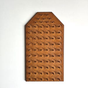 May include: A rectangular brown fiberboard pegboard with a pointed top edge. The board features a grid of circular indentations, each centered with a small wooden peg protruding outward. The surface has a textured, granular appearance throughout.