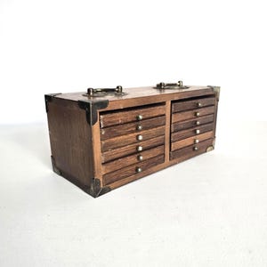 May include: A small, vintage wooden chest with multiple drawers. The chest is rectangular with metal corner reinforcements and small metal handles on top. Each drawer has a small, round metal knob. The wood has a warm, brown tone.