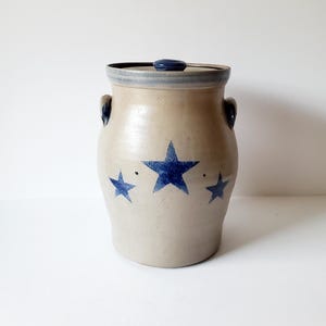 May include: A tan ceramic jar with a lid, featuring three blue stars. The jar has two handles and a blue rim. This vintage-style container is suitable for kitchen storage.