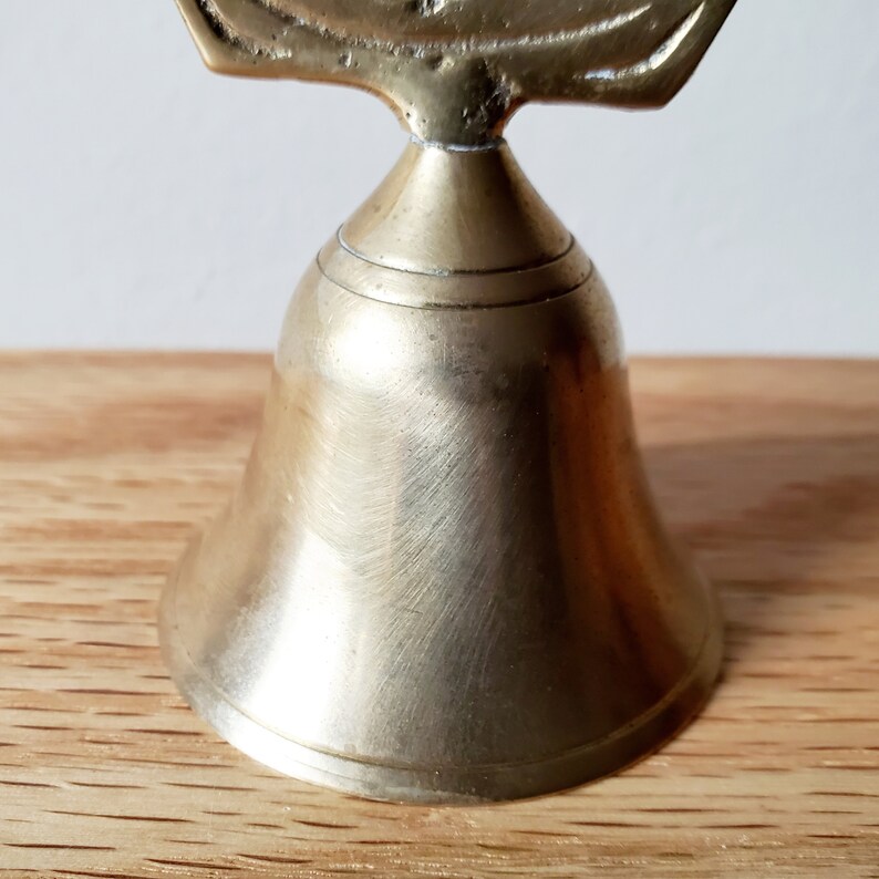 May include: A small, antique brass bell with a detailed handle. The bell has a classic shape with a flared base and a smooth, polished surface. The bell is sitting on a wooden surface.
