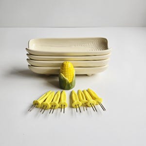 May include: A set of cream-colored rectangular serving dishes, a ceramic corn-shaped salt shaker, and yellow corn cob holders with metal prongs. The dishes are stacked, and the salt shaker is yellow and green.