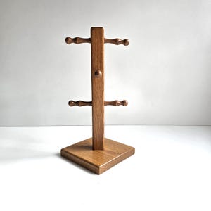 May include: A medium-brown wooden mug tree featuring a square base and a central vertical post. The post includes two sets of horizontal arms with rounded ends, designed for hanging mugs, and a small decorative knob centered between the arms.