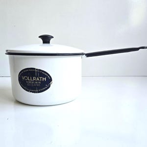 May include: A white enamel saucepan with a black rim, a black handle, and a matching lid with a black knob. A dark blue oval label on the side features the text VOLLRATH SINCE 1874, along with additional small printed text detailing the manufacturer.
