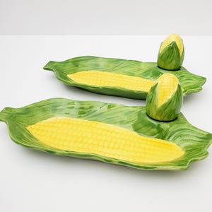 May include: Two ceramic corn-shaped serving dishes, each featuring a yellow cob resting on a green husk-shaped base. One dish includes a matching corn-shaped shaker piece attached to the end, while the other dish features a separate corn shaker.