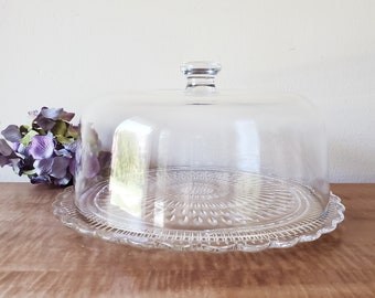 May include: A clear glass cake stand with a domed lid. The stand has a scalloped edge and a textured base.