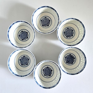 May include: Six white and blue porcelain bowls arranged in a circle. Each bowl features a central blue decorative emblem and a blue rim. The bowls have a delicate, openwork design around the sides.