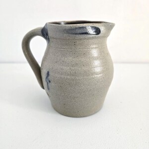 Op de afbeelding: Een steengoed kan met een handvat en een tuit. De kan is lichtgrijs van kleur met een donkerblauwe rand en handvataccenten. Een klein blauw ontwerp staat op de zijkant. De kan heeft een rustieke, handgemaakte uitstraling.