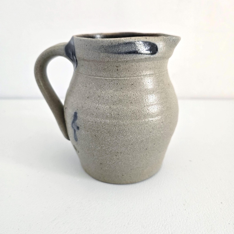 Op de afbeelding: Een steengoed kan met een handvat en een tuit. De kan is lichtgrijs van kleur met een donkerblauwe rand en handvataccenten. Een klein blauw ontwerp staat op de zijkant. De kan heeft een rustieke, handgemaakte uitstraling.