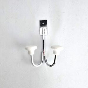 May include: A double wall hook with a polished chrome finish. The hook features two white, rounded knobs and a rectangular mounting plate. The hook is designed for hanging items and is mounted on a white wall.