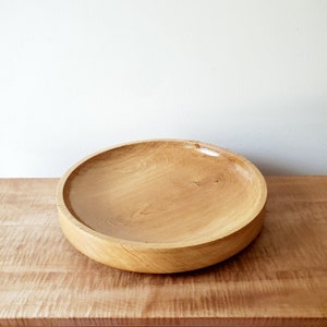 K&ouml;nnte beinhalten: Eine gro&szlig;e, runde Holzschale mit hellbraunem Finish. Die Schale ist aus Holz gefertigt und hat ein breites, flaches Design. Die Holzmaserung ist sichtbar und verleiht Textur. Die Schale steht auf einer Holzoberfl&auml;che.