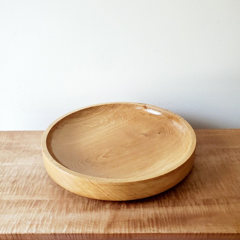 K&ouml;nnte beinhalten: Eine gro&szlig;e, runde Holzschale mit hellbraunem Finish. Die Schale ist aus Holz gefertigt und hat ein breites, flaches Design. Die Holzmaserung ist sichtbar und verleiht Textur. Die Schale steht auf einer Holzoberfl&auml;che.