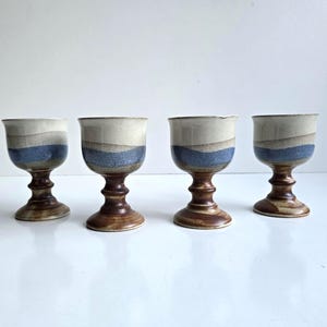 May include: Four ceramic goblets with a layered design. The top section is off-white, followed by a band of blue, and a band of tan. The base and stem are brown. The goblets are displayed on a white surface.