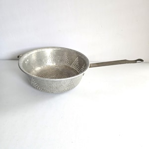 May include: A round, silver-colored metal colander with a long, flat handle extending from one side. The bowl features a perforated pattern of small holes arranged in triangular shapes throughout the basin and sides for draining.