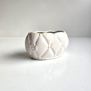 May include: An oval-shaped white ceramic planter featuring a raised diamond-quilted pattern across its exterior surface. Small, embossed flower motifs are positioned at the intersections of the quilted design, creating a textured, glossy finish.