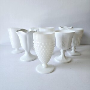 May include: A set of six white milk glass goblets with different designs. The goblets are all on a white surface.