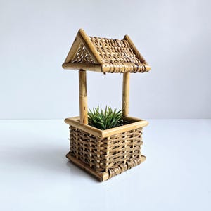 May include: A decorative miniature wishing well planter made of woven bamboo. The well features a square base, vertical supports, and a sloped roof. A green plant sits inside the well. The planter is a natural light brown color.