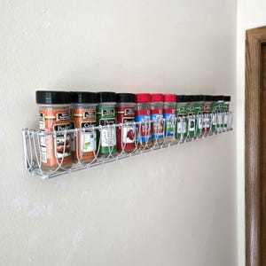 May include: A white wire spice rack mounted on a wall, holding a row of twelve clear plastic spice jars with black lids. Each jar features a label with text including Spice Supreme, Poultry Seasoning, Celery Salt, Thyme Leaves, and Paprika.