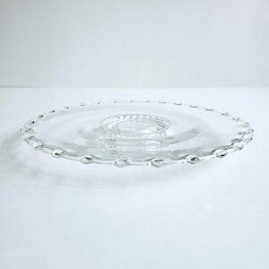 May include: A clear glass serving plate with a beaded edge. The plate has a raised, circular center and a wide, flat surface. The edge is adorned with small, evenly spaced, rounded beads. The plate is designed for serving food.