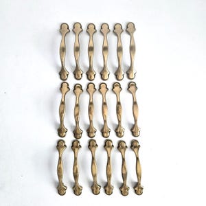 May include: A collection of twenty-one antique brass-toned cabinet pulls. Each pull features a decorative, elongated design with a central handle and ornate ends. The pulls are arranged in a grid pattern against a white background.