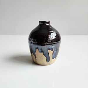 May include: A handmade ceramic vase with a dark brown, glossy top and a light tan base. The vase has a unique design with dark blue glaze dripping down the sides. The vase is a decorative item.