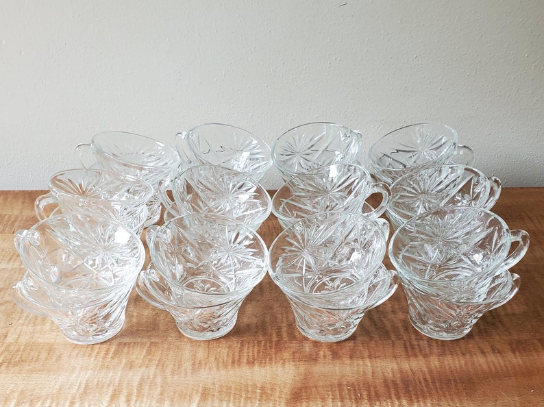 May include: A set of 12 clear glass cups with a starburst pattern. The cups are stacked in two rows of six, with the top row slightly offset from the bottom row.