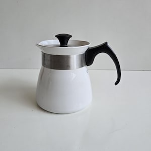 May include: A white coffee pot with a black handle and lid. The pot has a silver band around the middle and a curved handle. The pot is sitting on a white surface.