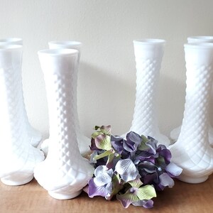 Puede incluir: Conjunto de ocho jarrones de vidrio de leche blanco con un patr&oacute;n de diamante. Los jarrones est&aacute;n dispuestos en una fila con un ramo de flores moradas y verdes delante de ellos.