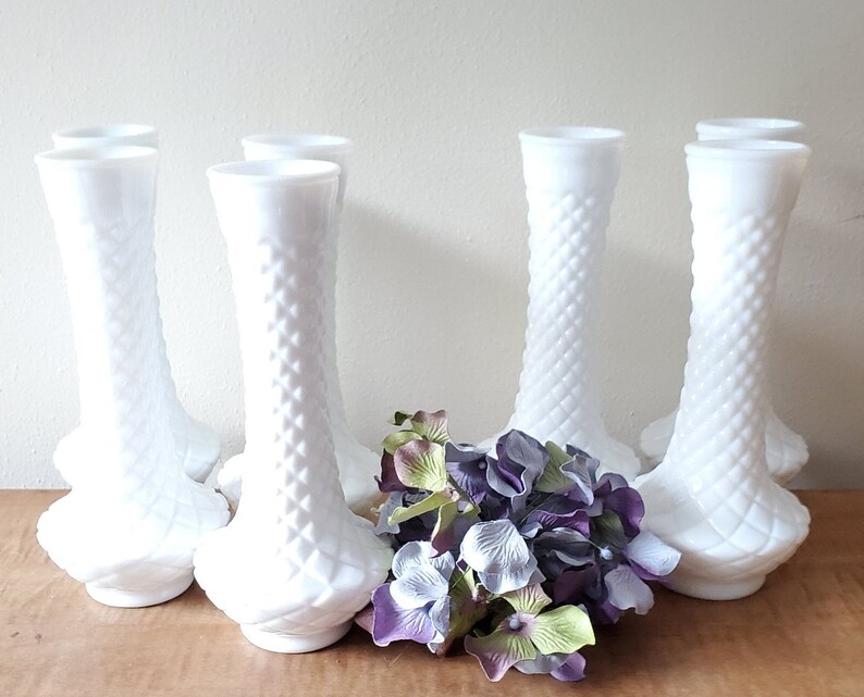 Puede incluir: Conjunto de ocho jarrones de vidrio de leche blanco con un patr&oacute;n de diamante. Los jarrones est&aacute;n dispuestos en una fila con un ramo de flores moradas y verdes delante de ellos.