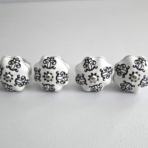 May include: Four white ceramic cabinet knobs with a black floral design and a silver center. The knobs are arranged in a row and have a scalloped edge. These decorative knobs are suitable for furniture.