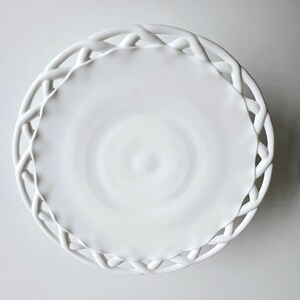 May include: White ceramic cake stand with a woven edge design. The stand is round and has a smooth, white surface.