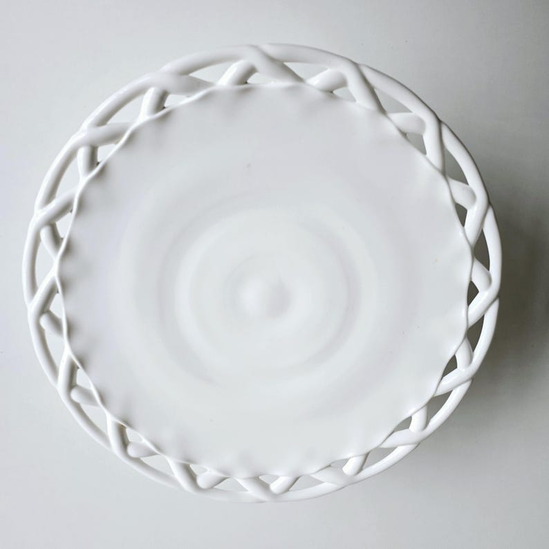 May include: White ceramic cake stand with a woven edge design. The stand is round and has a smooth, white surface.